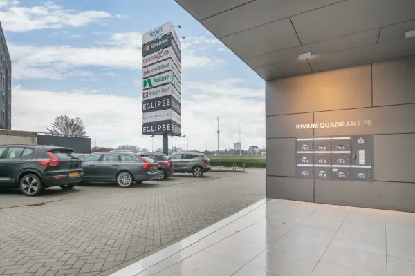 Ingang en parkeerplaats bij kantoorgebouw Rivium Quadrant 75 met bedrijfsnaambord op de achtergrond.